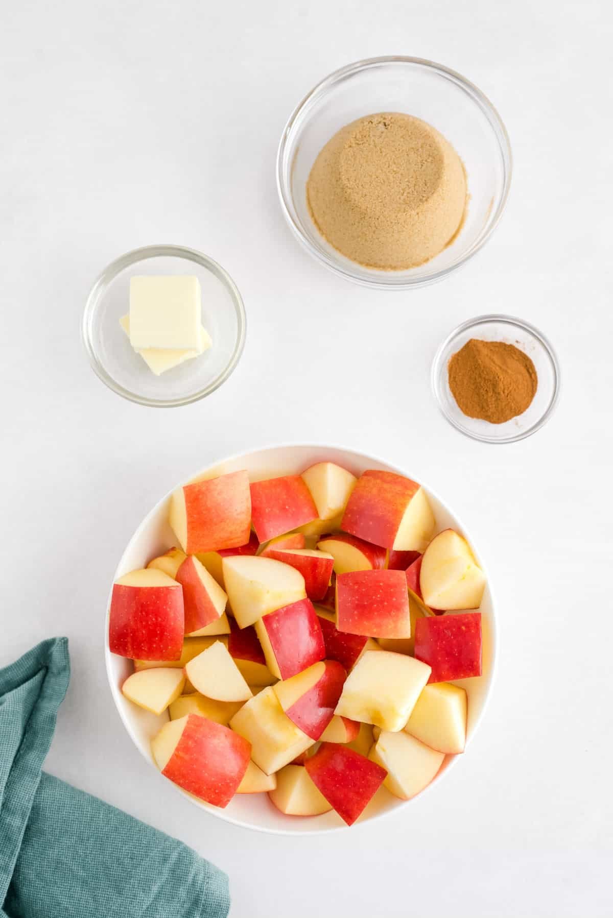 Quatro tigelas cheias de ingredientes: maçãs picadas, manteiga, açúcar mascavo e canela.