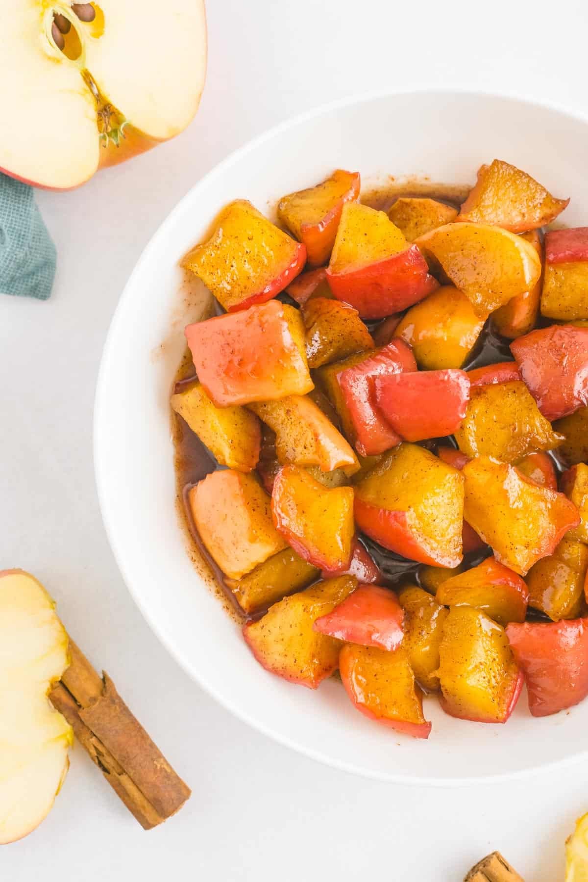 Compota de maçã e canela em uma tigela branca, com metades de maçã e paus de canela também na foto.