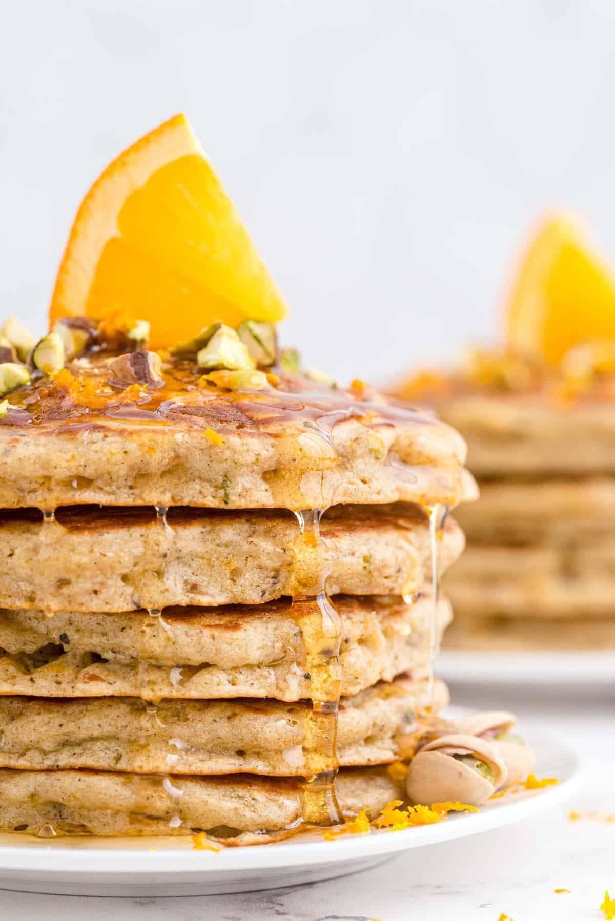 Pilha de panquecas num prato branco, cobertas com pistaches, mel e uma fatia de laranja.