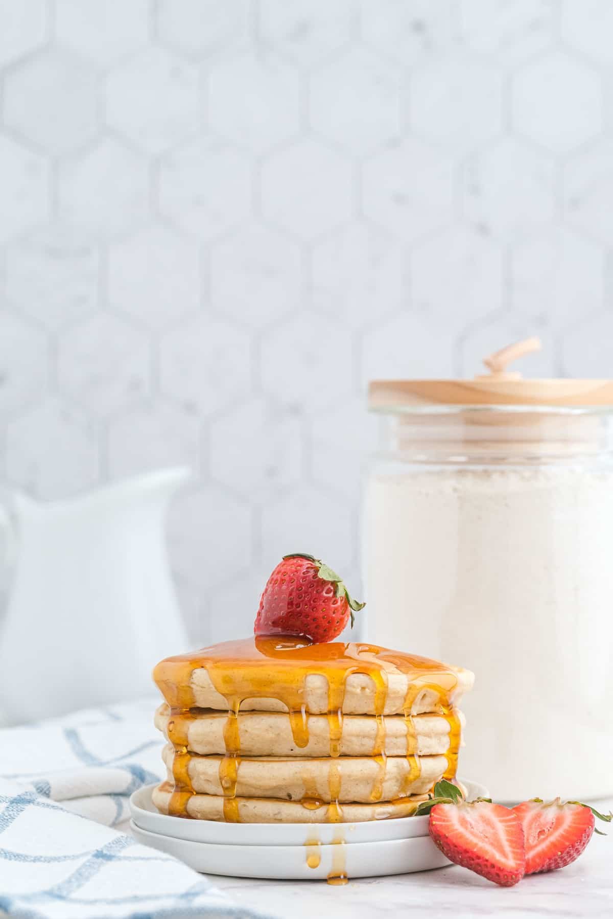 Pilha de panquecas com xarope e morangos, um recipiente de mistura de panqueca ao fundo.