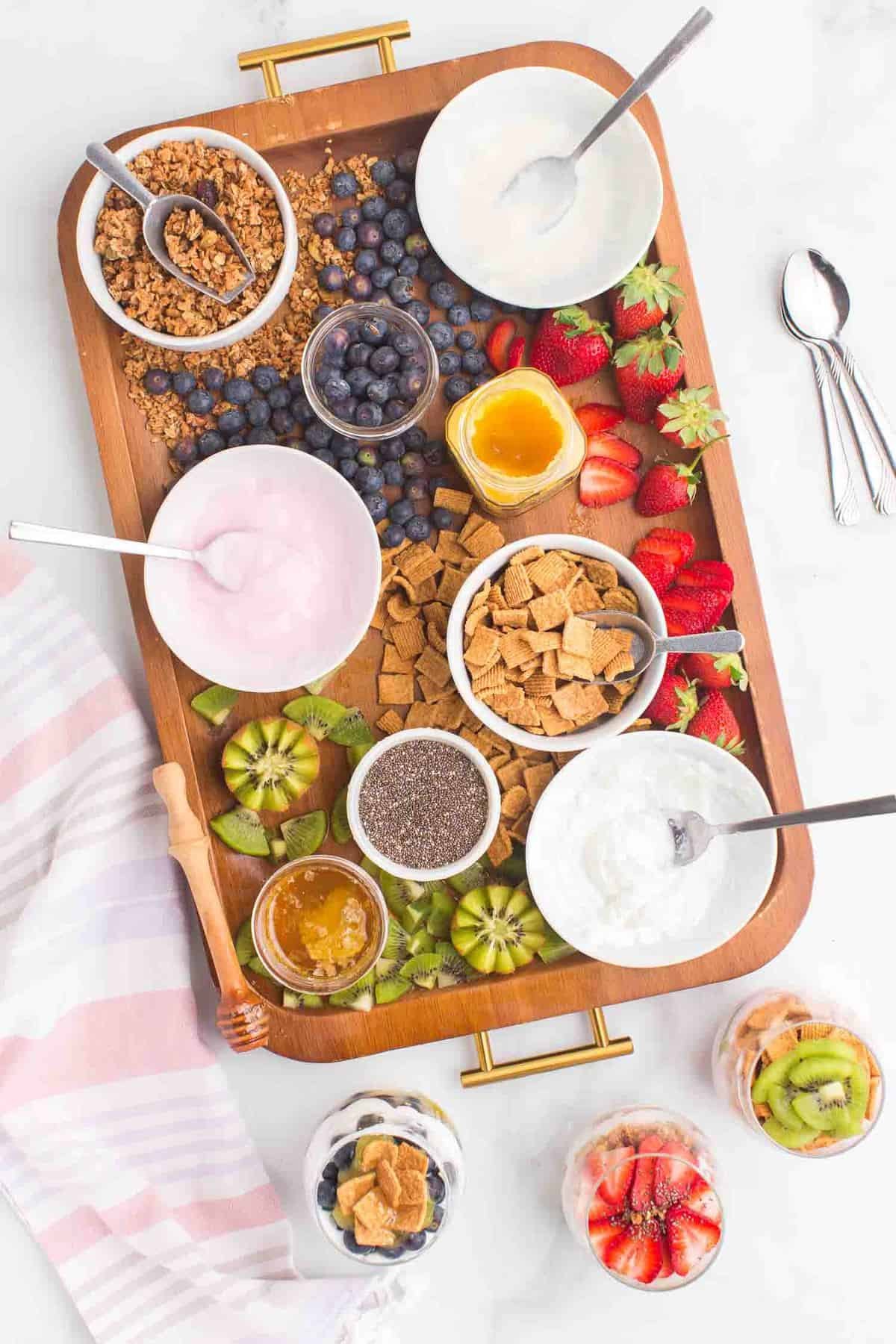 Iogurte e acompanhamentos dispostos em uma bandeja, três parfaits ao lado.