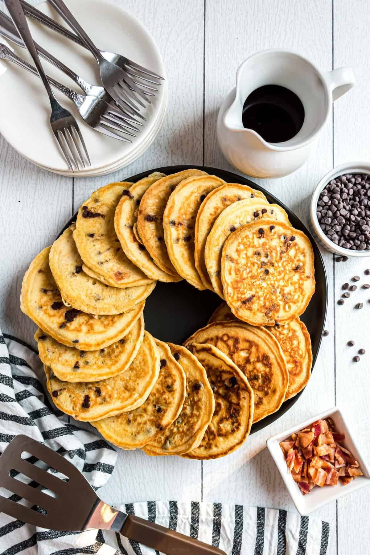 Prato redondo preto cheio de panquecas de bacon com chocolate, garfos e pratos próximos.