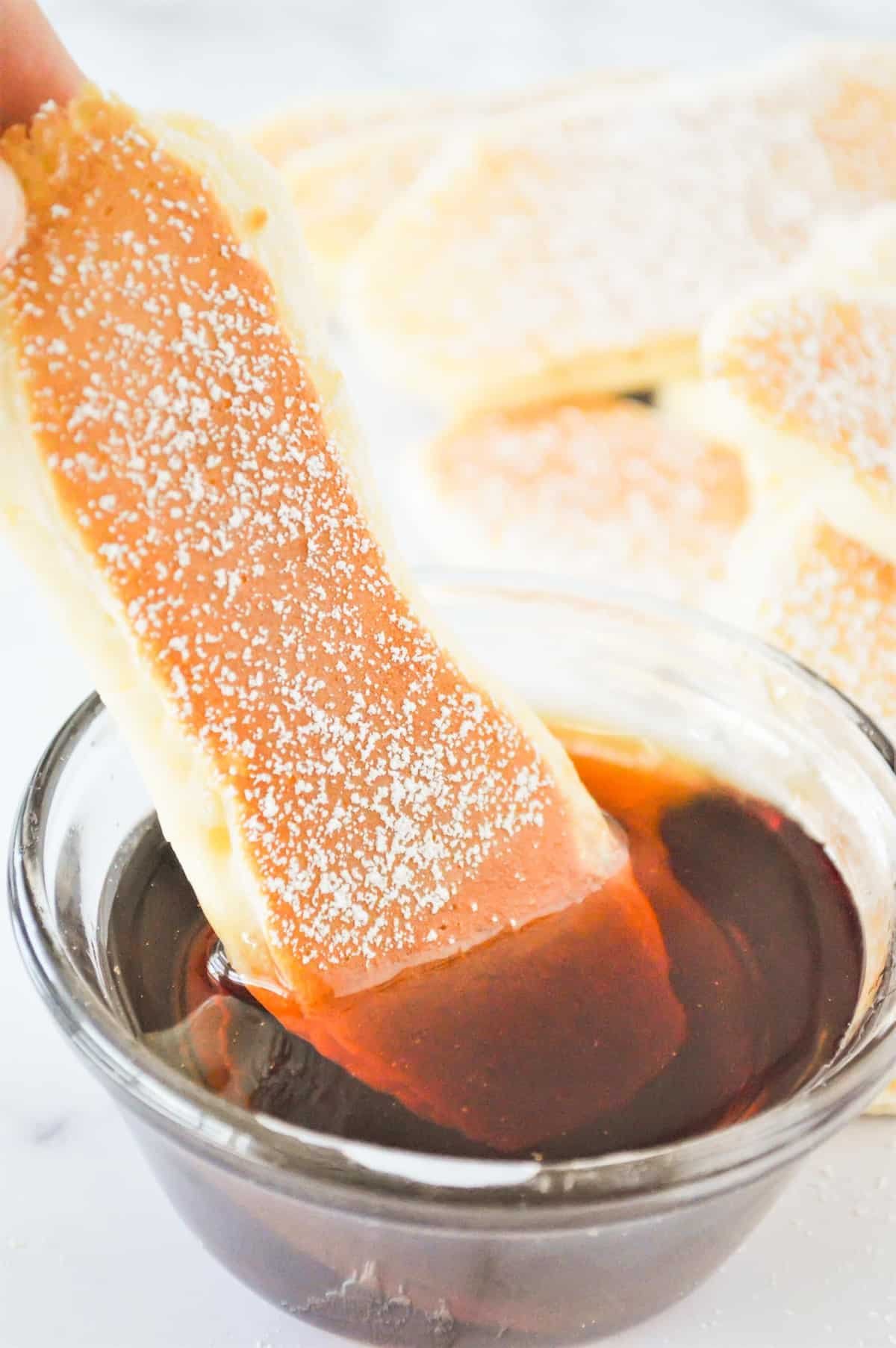 Panqueca fina e comprida sendo mergulhada em uma tigela de xarope.