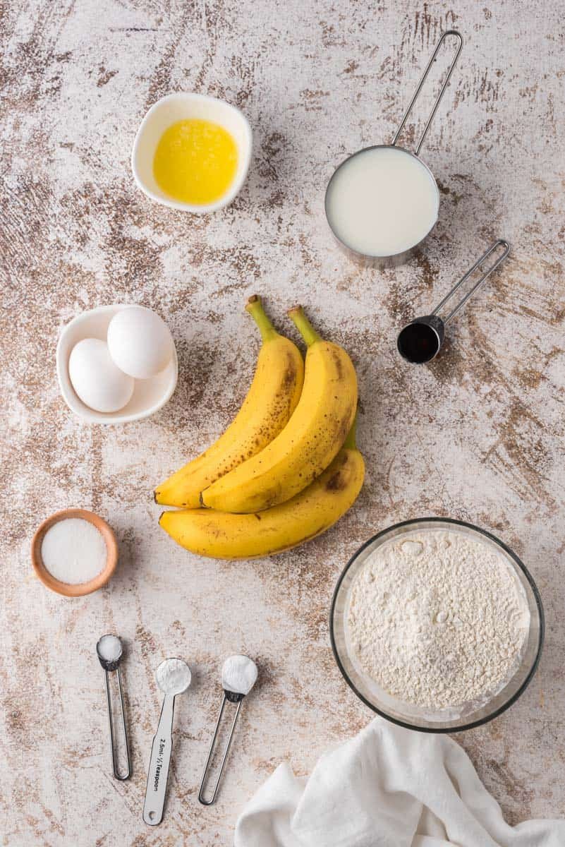 Ingredientes em uma superfície marrom e branca: farinhas, açúcares, bananas maduras, ovos, óleo, extrato de baunilha.