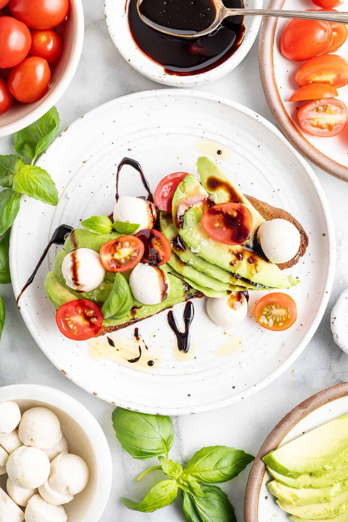 Toast de abacate coberto com tomates e mussarela, cercado por ingredientes adicionais.