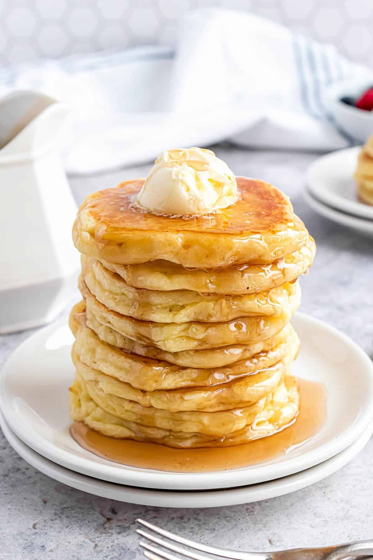 Pilha alta de panquecas num prato branco, com manteiga e xarope.