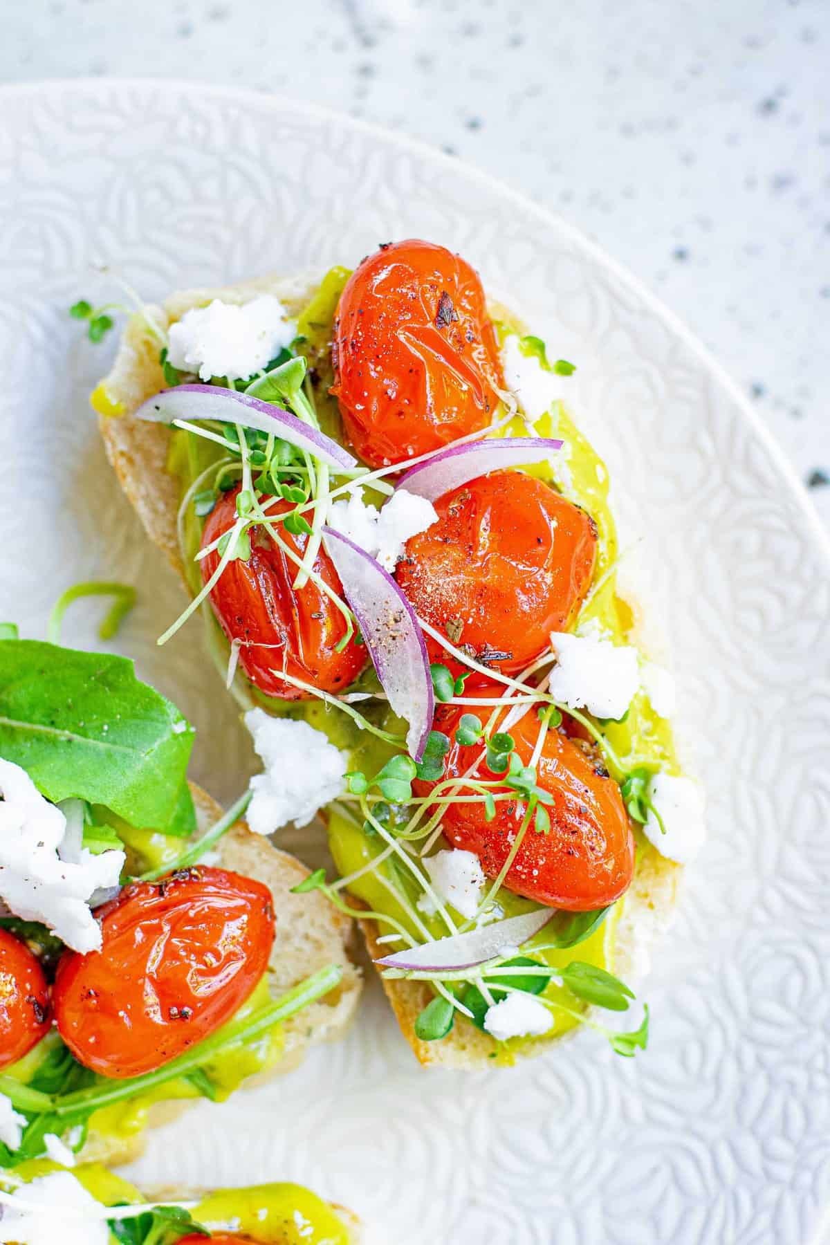 Vista aérea de pequeno pedaço de pão coberto com guacamole, tomates assados, folhas verdes e feta.