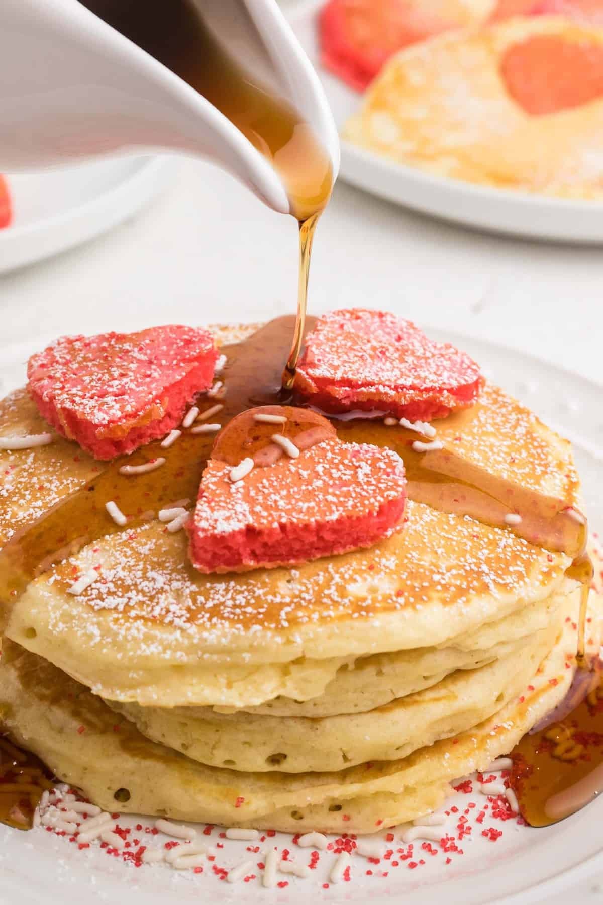 Xarope sendo derramado sobre panquecas com mais panquecas em forma de coração em cima.