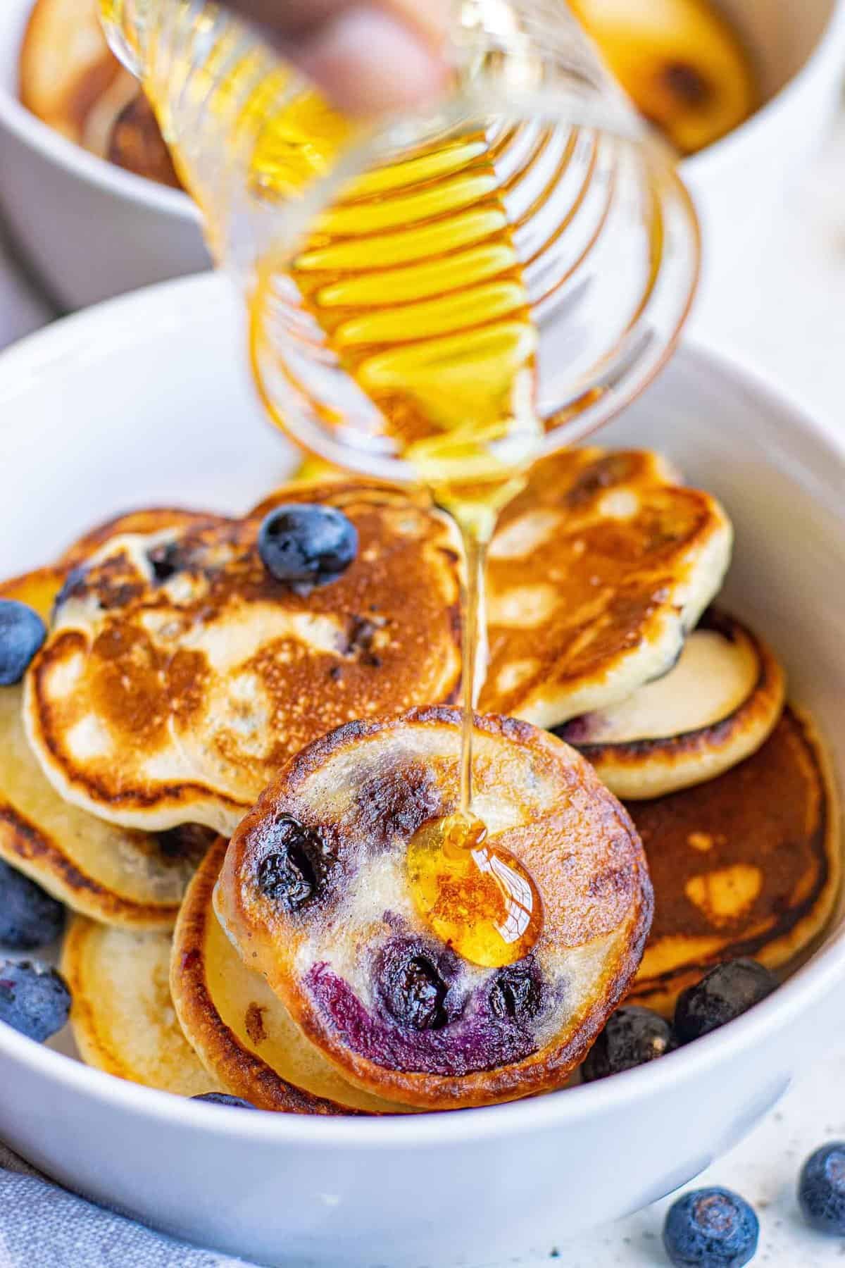 Xarope sendo derramado em uma tigela de mini panquecas de mirtilo.