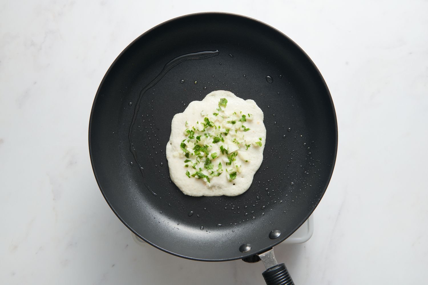 A greased nonstick skillet with a small portion of batter in the center, topped with chopped onions, chiles, and cilantro