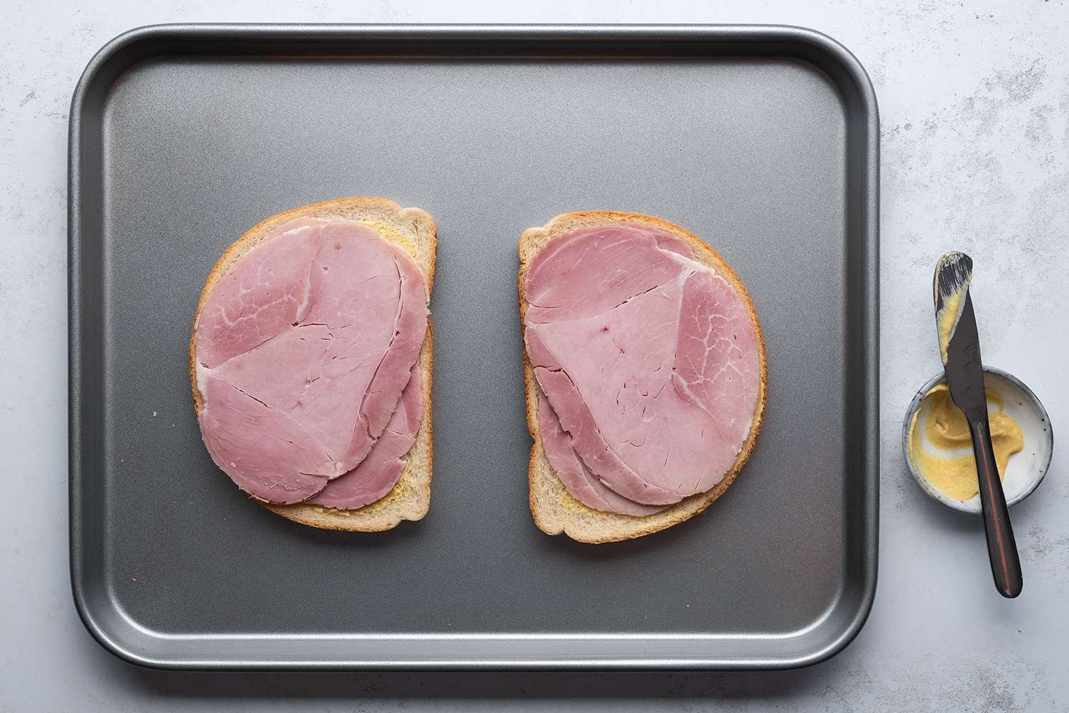 Bread slices with mustard and ham on a sheet pan, and a knife on top of a bowl with mustard