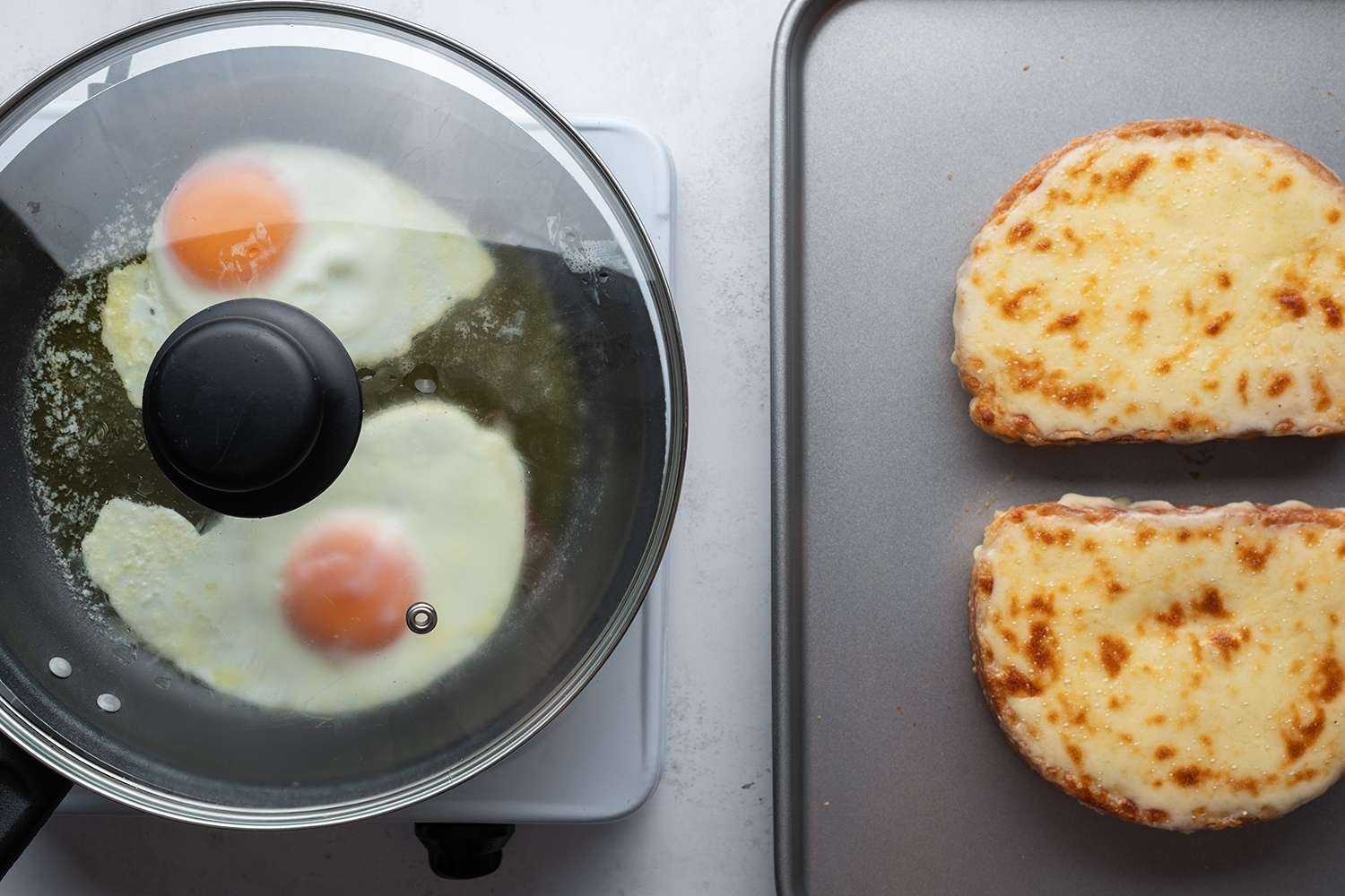 Eggs cooking in a covered pan on a burner, and two sandwiches on a sheet pan