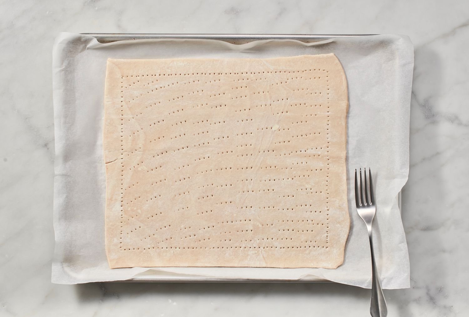 A rectangular piece of puff pastry on a parchment paper-lined baking sheet covered with fork prick marks