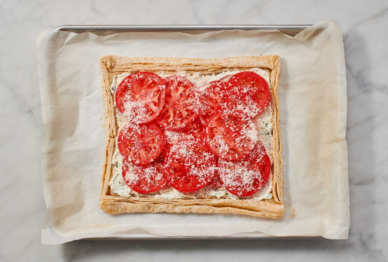 A partially baked puff pastry crust topped with cream cheese mixture and sliced tomatoes, topped with cheese and black pepper